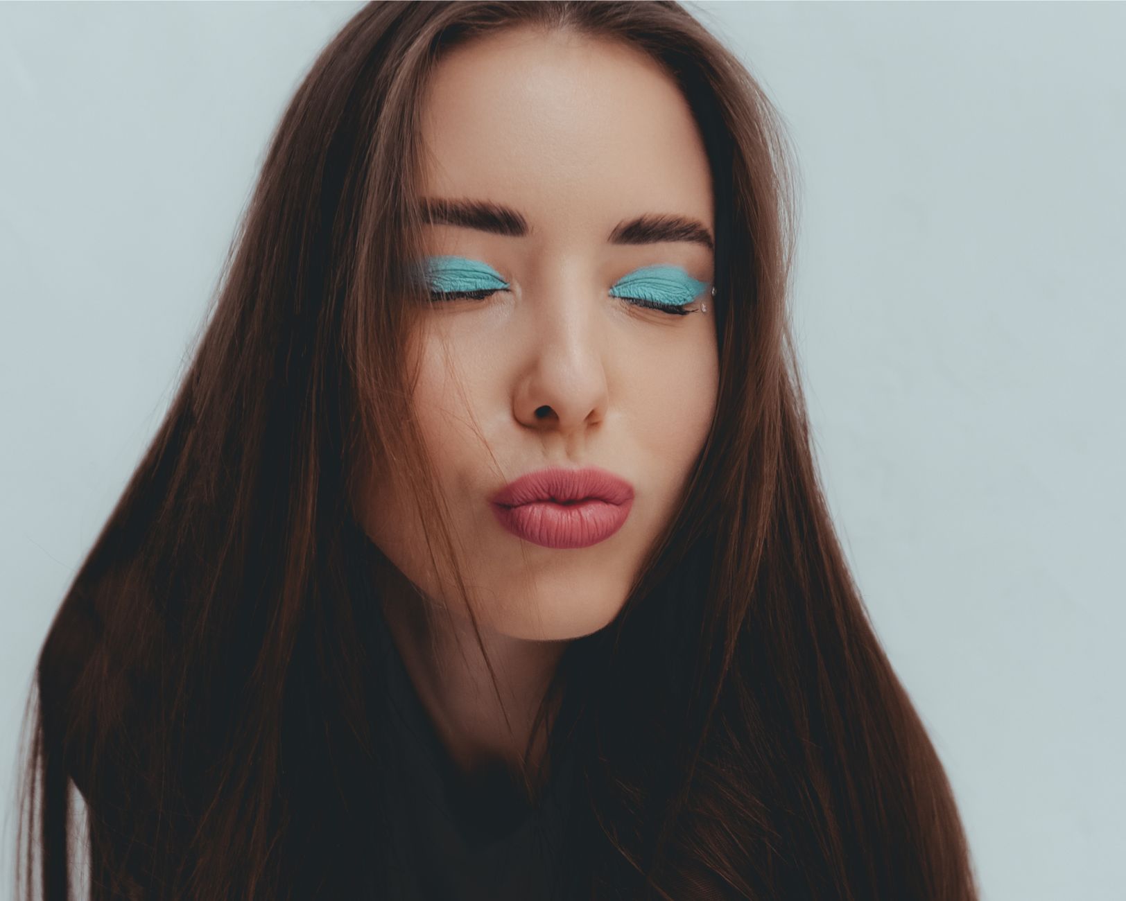 Studio portrait of a woman with vibrant blue eye shadow and pouting lips, eyes closed.