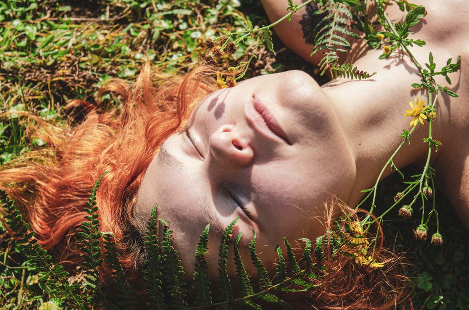A woman with red hair relaxes amidst ferns and yellow flowers under the sun, experiencing nature's tranquility.