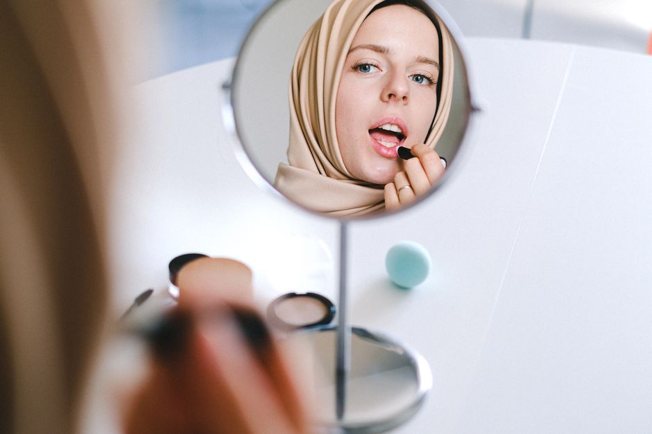 Muslim woman in hijab applies lipstick while looking at her reflection in a mirror.
