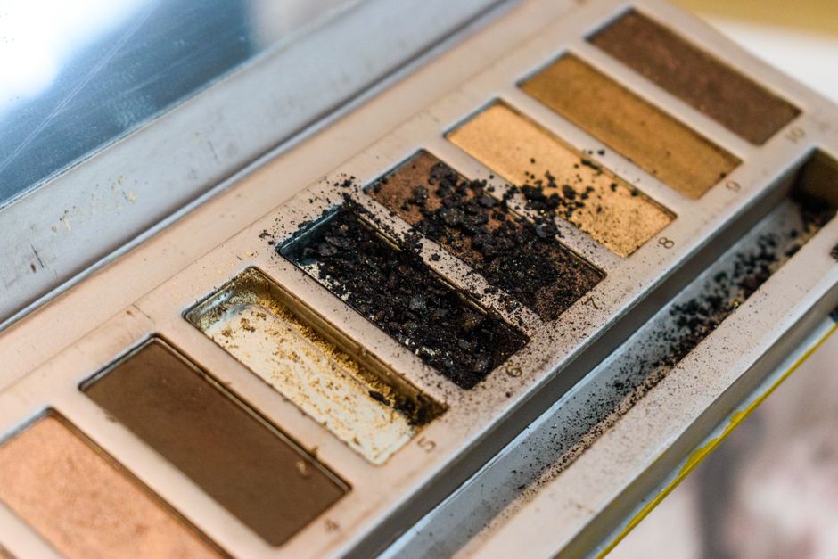 A detailed close-up of a makeup eyeshadow palette with visible powder and dust, highlighting a range of neutral colors.