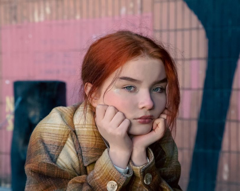 Close-up portrait of a pensive redheaded teenage girl outdoors, leaning on her hands, wearing a checkered coat.