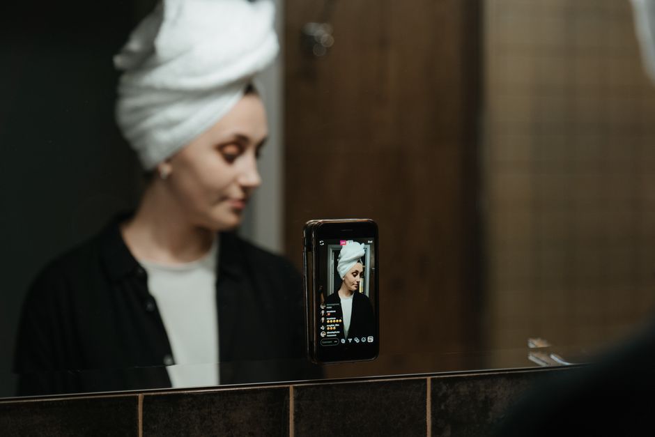 A woman reflects in a mirror during her beauty routine with a towel on her head, using a smartphone.
